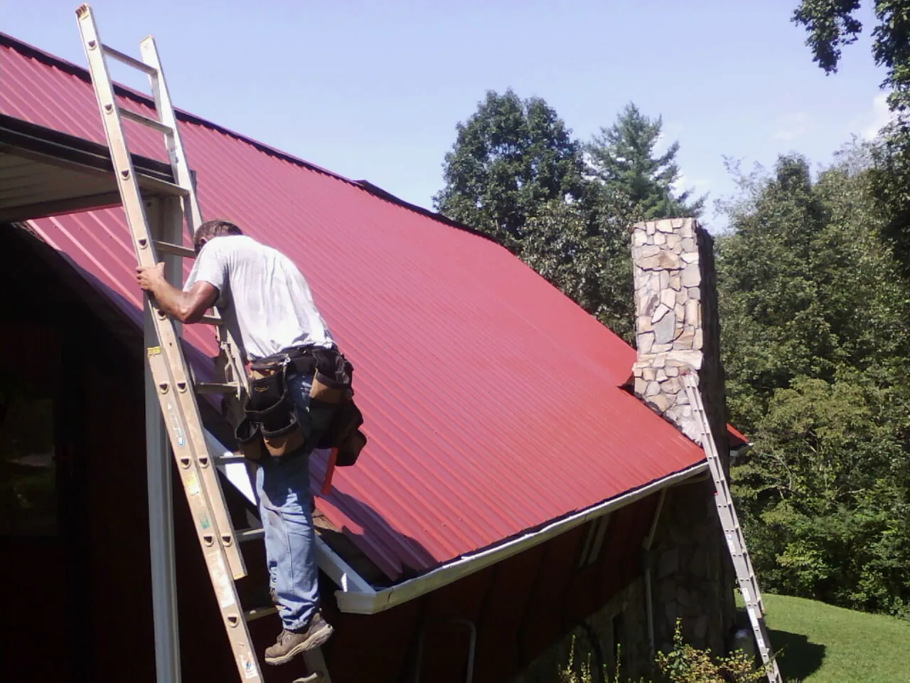 Roofer on a ladder beside a red metal roof and stone chimney — Soffit & Fascia Repair by Roofing Brothers in Abbeville